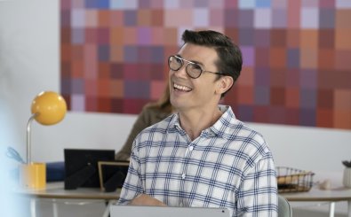 Ryan O’Connell as Ryan Hayes, a male with brown hair and eyeglasses, wears a blue and white patterned button down and a smile on his face. He is seated at a desk and a vibrant office is visible behind him. Bild: Cr. Beth Dubber / Netflix (C) 2021