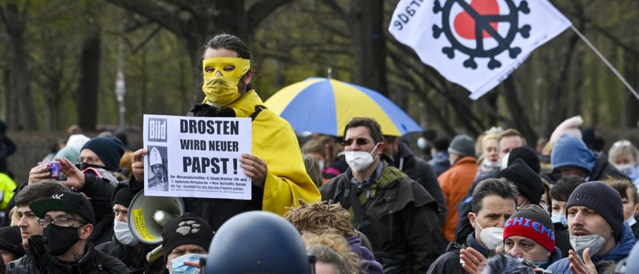Police observe a masked protestor taking part in a demonstration holding a poster with an image of German virologist Christian Drosden reading, „Drosden will be the new Pope“ in Berlin on April, 13, 2021, as anti lockdown critics and so-called ‚Querdenker‘ gathered outside the German parliament on Tuesday, amid the Covid-19, corona pandemic. (Photo by John MACDOUGALL / AFP)