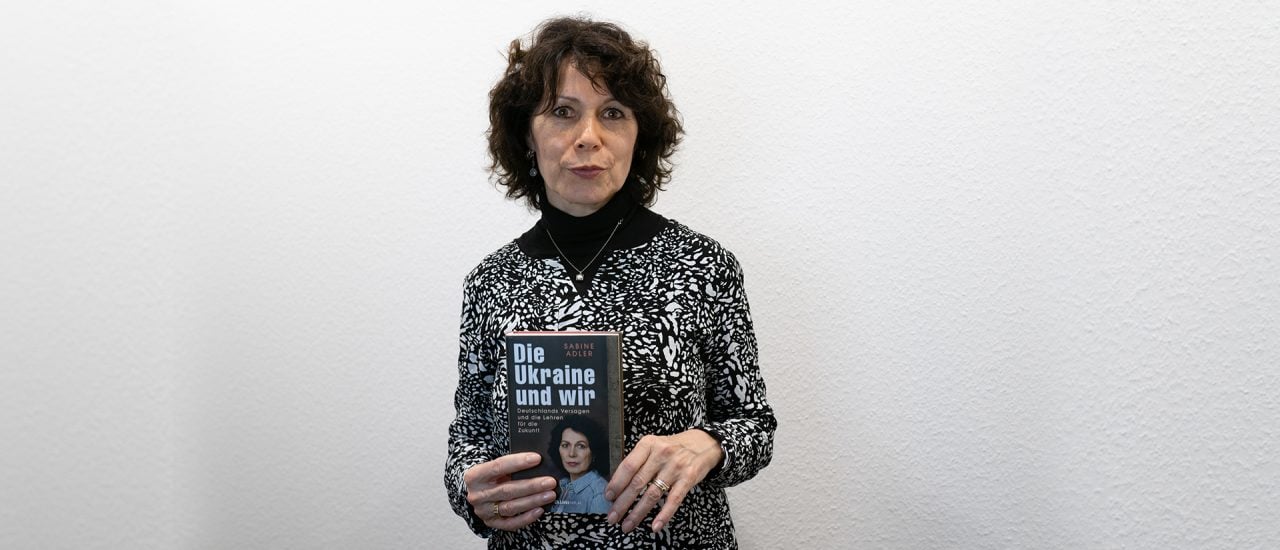 Sabine Adler vor einer weißen Wand. In ihrer Hand hält sie ihr aktuelles Buch.