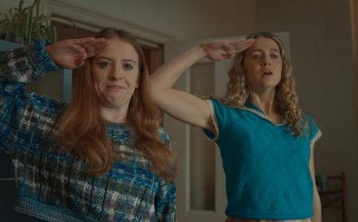 Die rothaarige Jen (links) steht neben ihrer blonden Freundin Carrie (rechts). Beide salutieren mit der rechten Hand an der Schläfe. Jen ist kurz davor loszulachen, Carrie schaut ernster. 