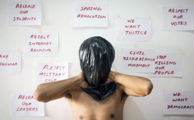 Eine männlich gelesene Person steht ohne Shirt und mit einer schwarzen Plastiktüte auf dem Kopf vor einer weißen Wand. Auf der Wand sind in roter Schrift auf Papier Forderungen zu lesen wie „We want justice“.