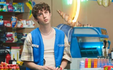 Der lockige Caleb trägt ein helles Shirt mit einer blauen Weste, eine Arbeitsuniform, darüber. Er sitzt an einer Supermarktkasse und schaut gelangweilt in die Gegend. 