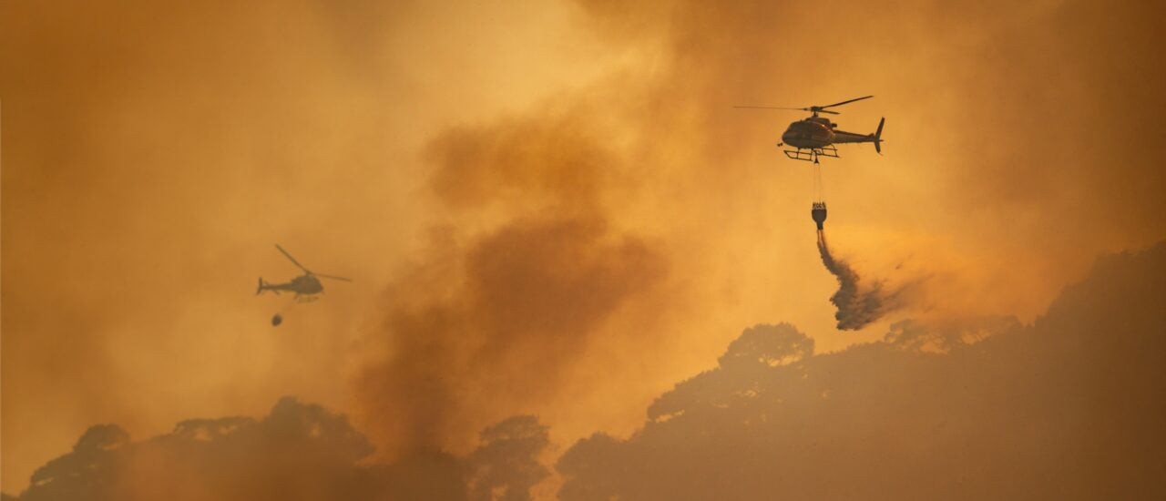 Zwei Löschhubschrauber im Einsatz, sie fliegen über einen orange gefärbten, von Rauchwolken geschwängerten Himmel.