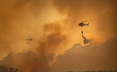 Zwei Löschhubschrauber im Einsatz, sie fliegen über einen orange gefärbten, von Rauchwolken geschwängerten Himmel.