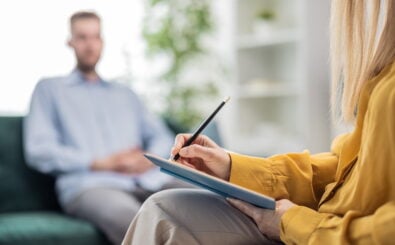 Symbolbild Psychotherapie: Ein Mann (unscharf) sitzt auf einem Sofa. Davor eine Frau mit einem Klemmbrett in der Hand. Foto: Shutterstock/MYDAYcontent