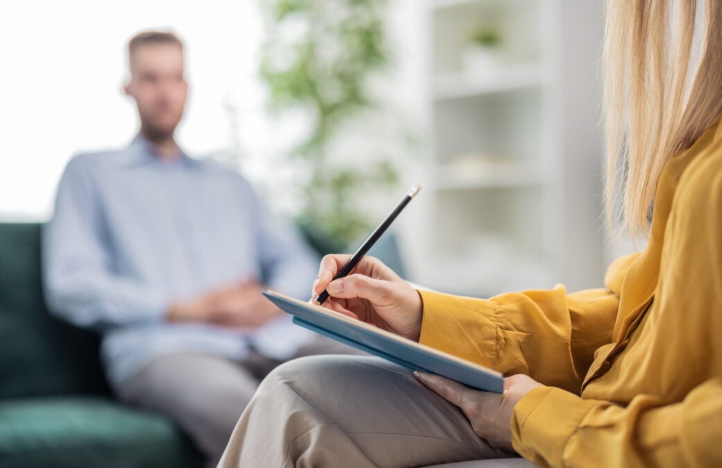 Ein Mann (unscharf) sitzt auf einem Sofa. Davor eine Frau mit einem Klemmbrett in der Hand. Foto: Shutterstock/MYDAYcontent